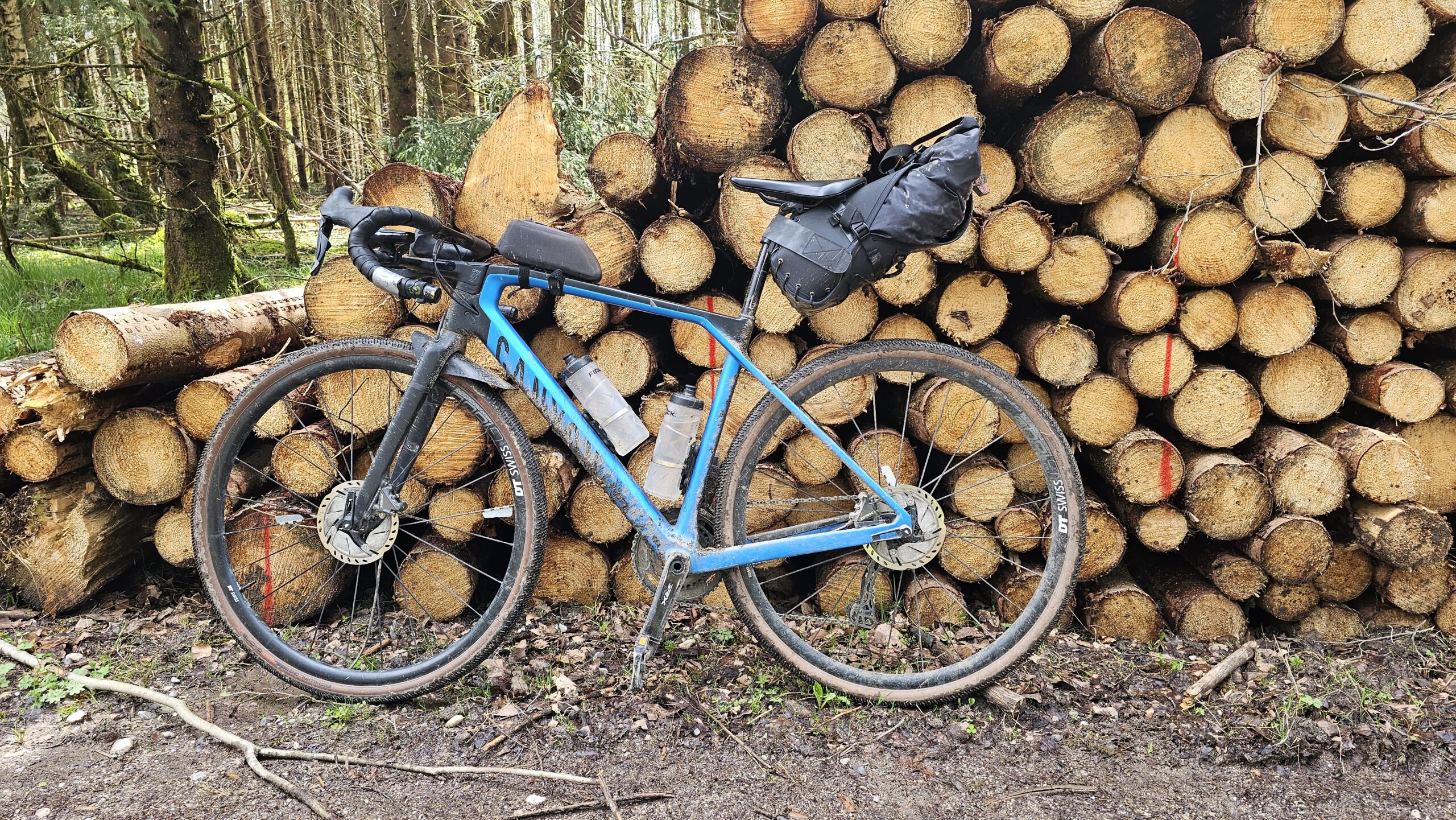 Hellblaues Gravelbike mit Satteltasche vor einem Holzstapel im Wald